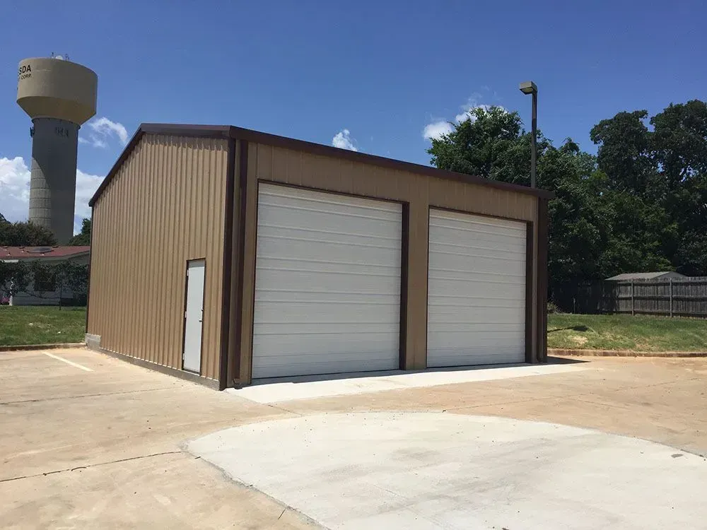 A tan, metal-sided commercial garage with two white roll-up doors and a side entry door, located near a water tower.