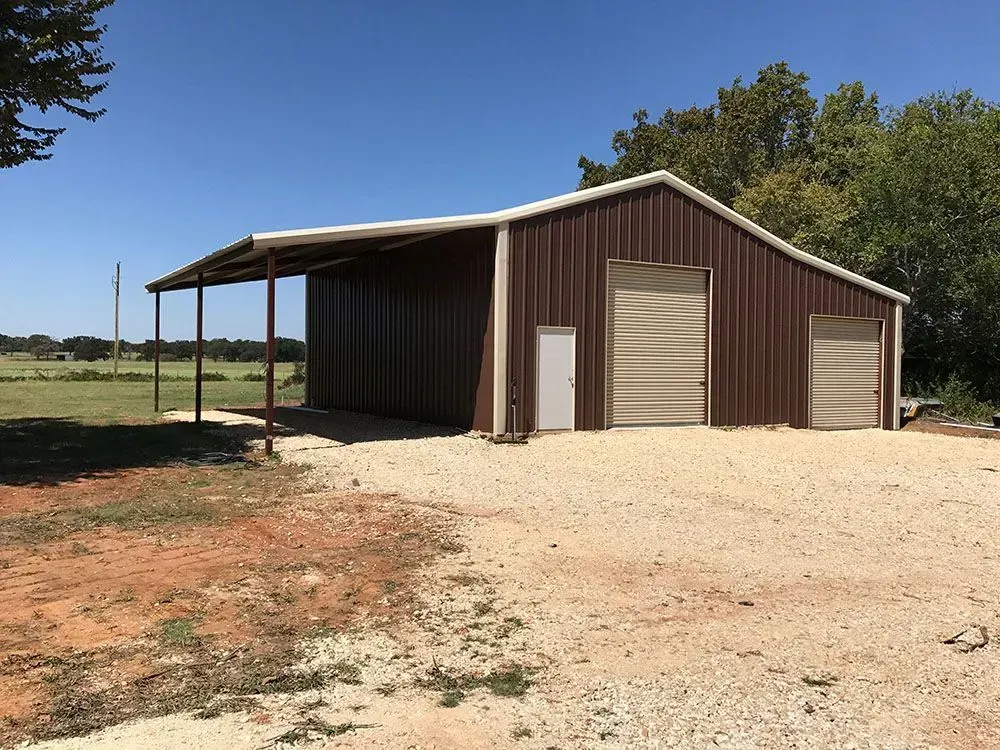 A dark brown metal barn with a roll-up garage door, a side entry door, and an open-sided carport on a gravel lot.