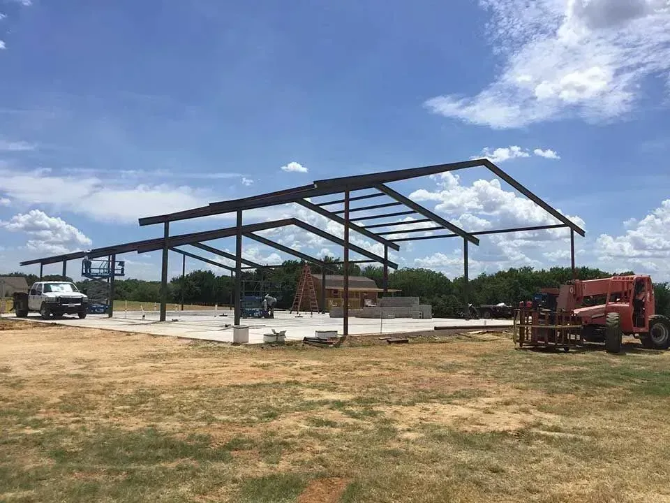 A steel frame structure for a building under construction on a concrete foundation in an open field under a blue sky.