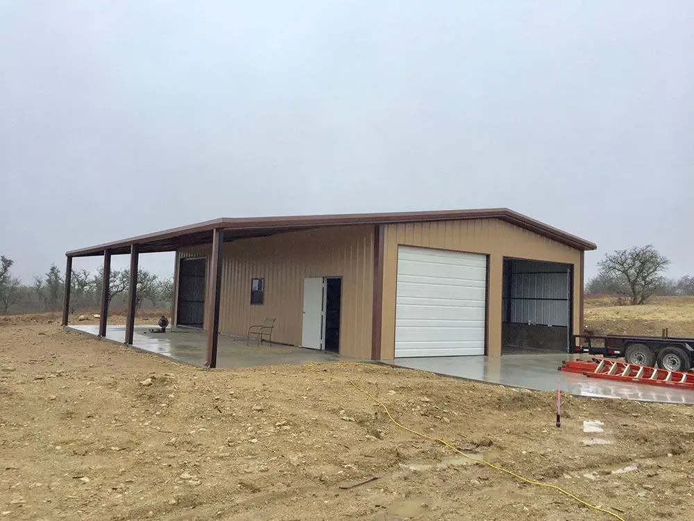 A tan metal workshop with a side carport, two garage bays, and a side entry door, set on a dirt lot under a cloudy sky.