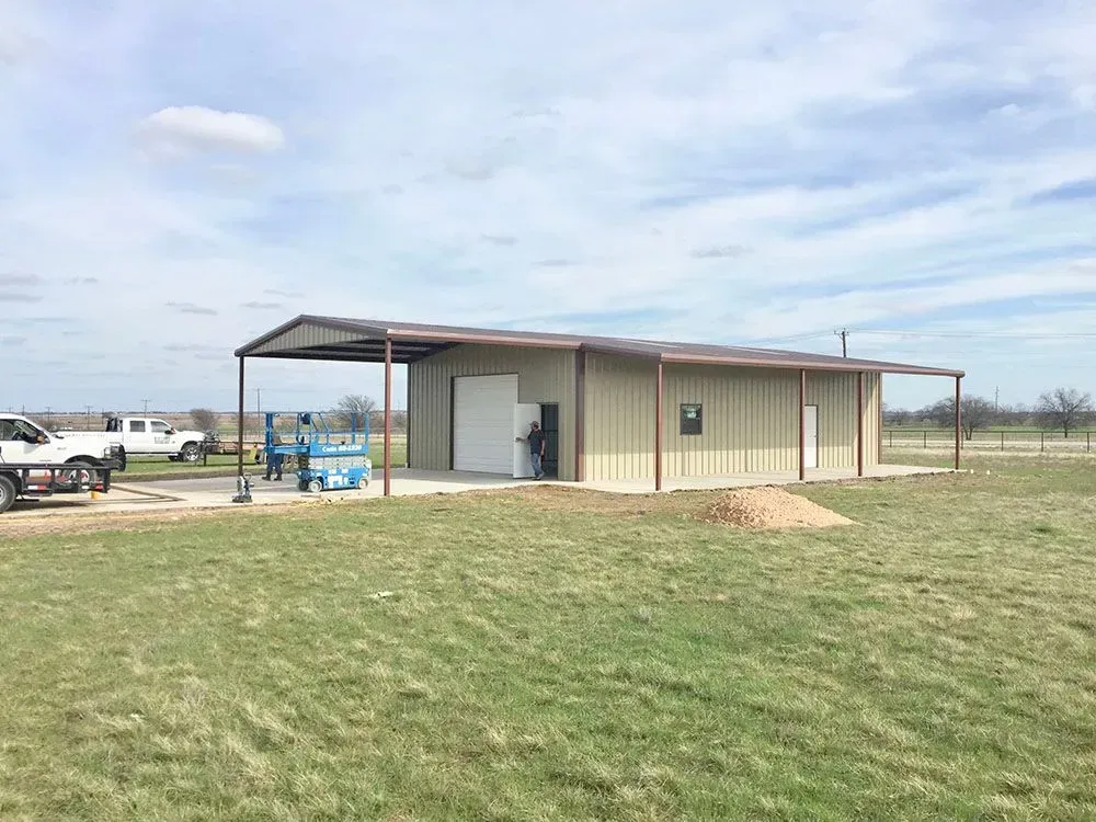 A metal garage with a covered patio, parked trucks, and workers under a blue sky in an open, grassy field.