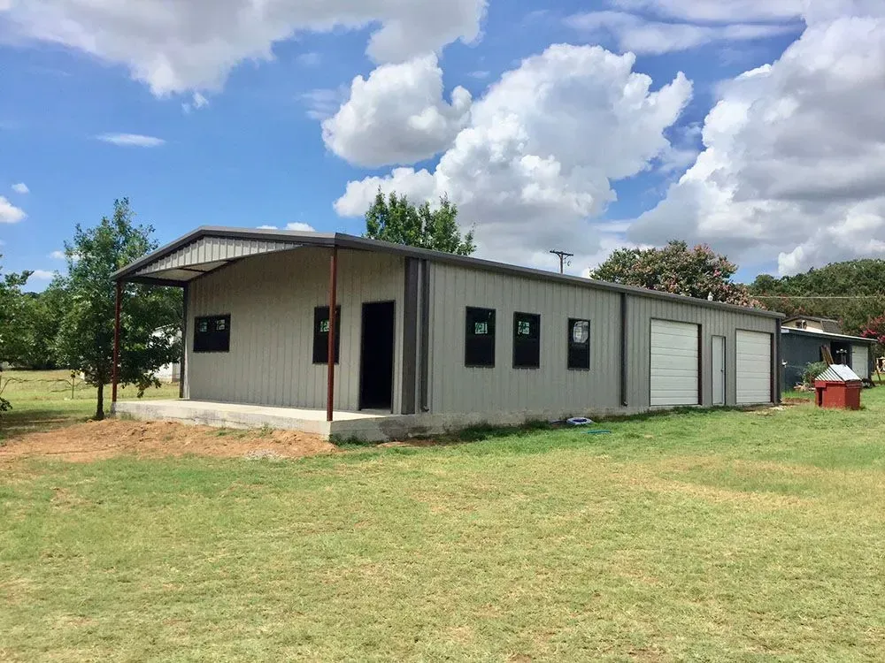A beige metal-sided building with a covered front porch, garage doors, and windows, set in a grassy field under a blue sky.