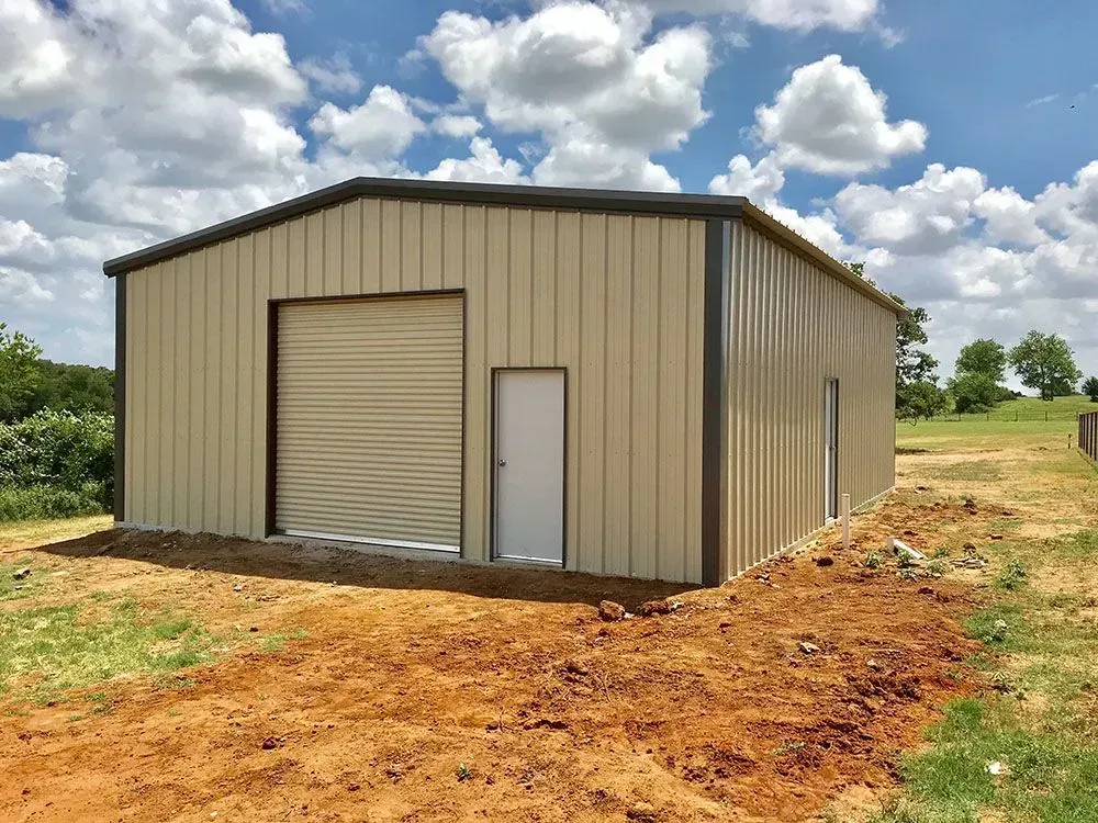 A tan metal-sided garage with a brown trim, a roll-up door, and a side entry door, set in a grassy field under a blue sky.