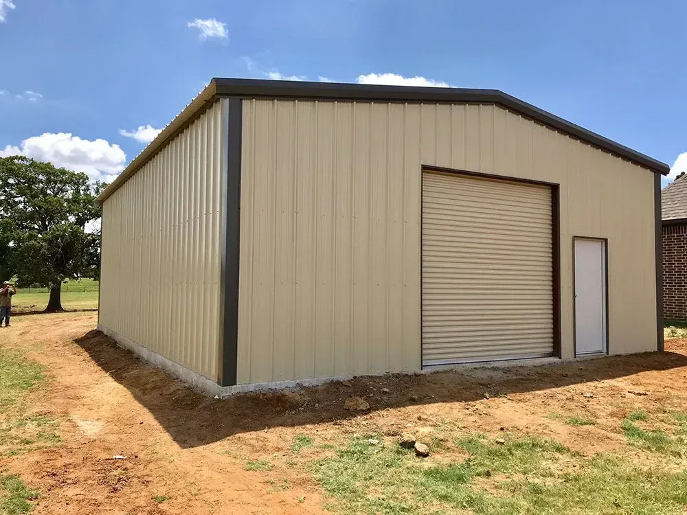 A beige metal warehouse with a roll-up garage door and side door sits on a dirt lot under a blue sky.