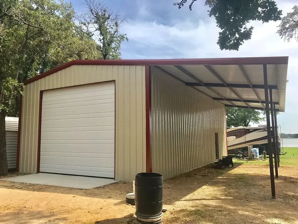 A tan metal garage with a white roll-up door and a side carport, set in a rural, wooded area.