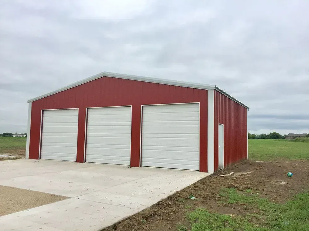 A red metal building with three white garage doors and a side entry door, set on a concrete pad in a grassy field.