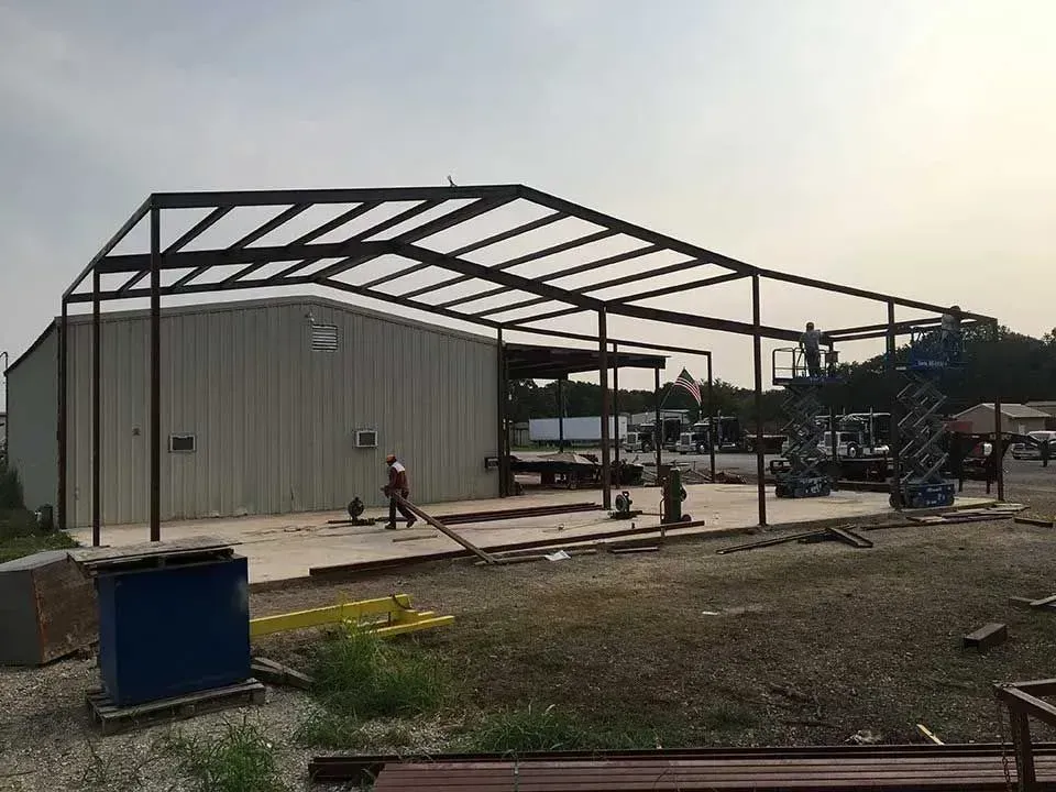 Workers assemble a metal-framed structure extension attached to an existing industrial building.
