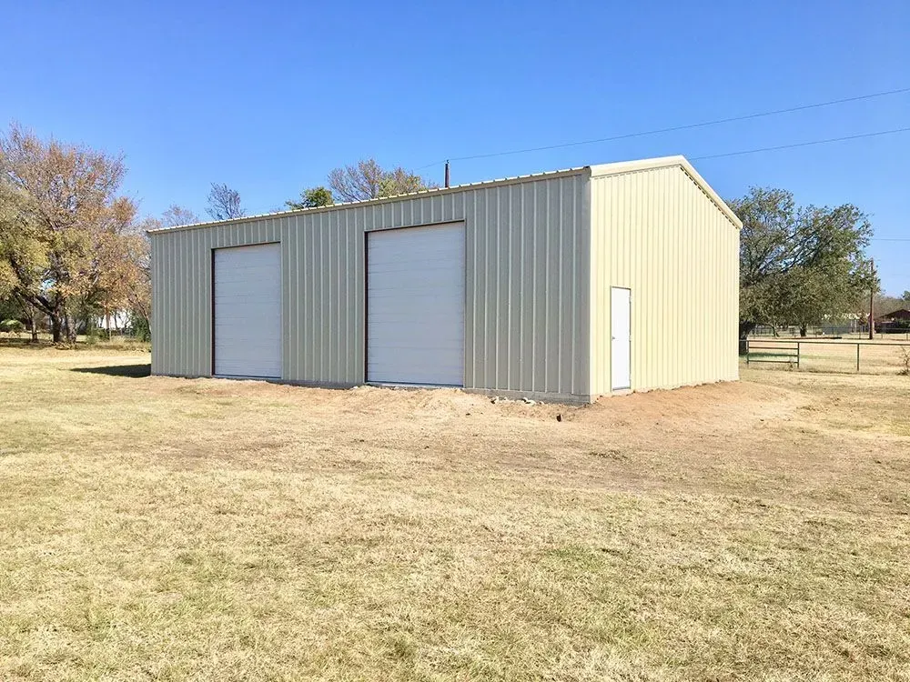 A tan metal workshop with two garage doors and a side entry door, situated in a dry, grassy field under a clear blue sky.
