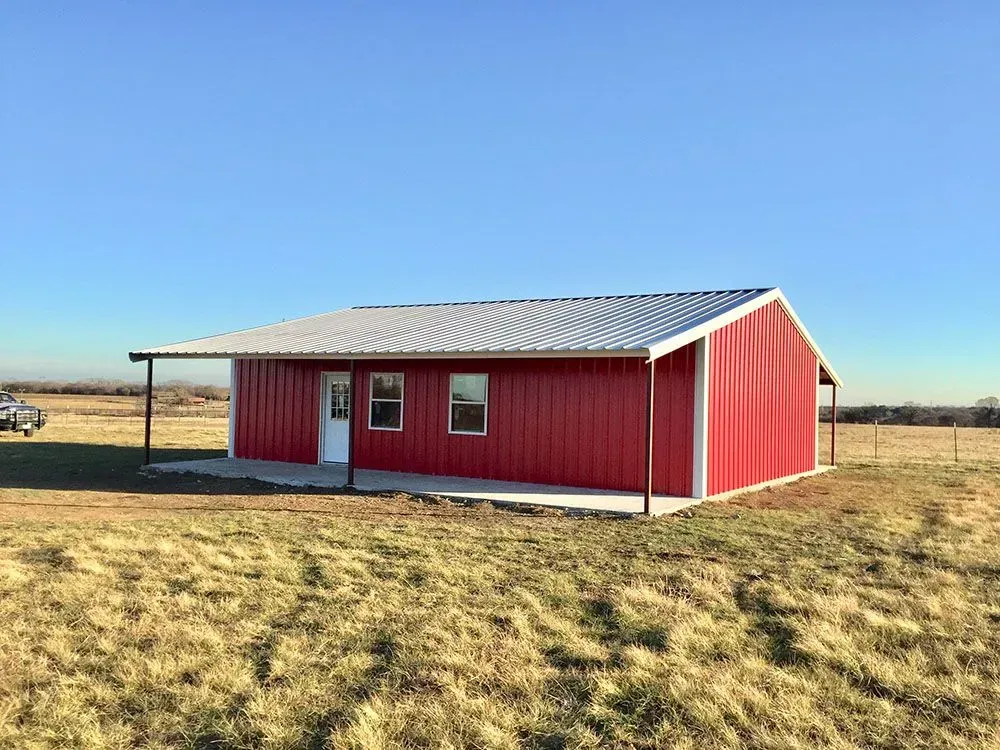 A red metal building with a white roof and a front porch sits in a large, open field under a clear blue sky.