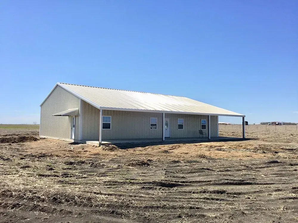 A tan, metal-sided barndominium-style house with a front porch sits on a flat, dirt lot under a clear blue sky.