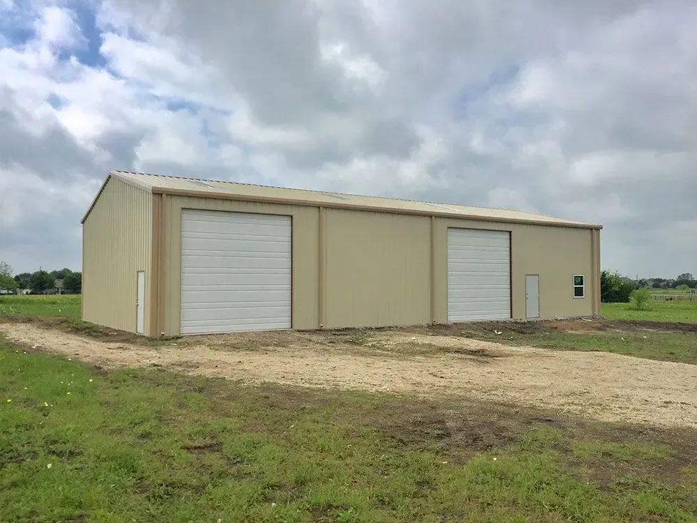 A tan, metal workshop building with two white roll-up doors, set in a grassy field under a cloudy sky.