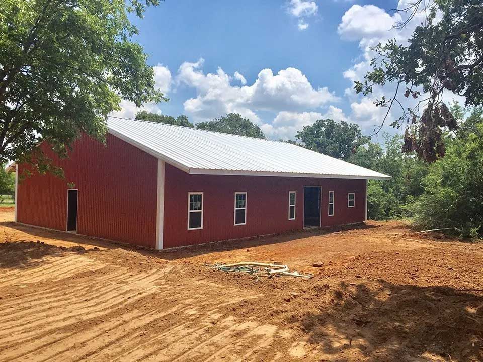 A newly constructed red metal building with a white roof stands on a cleared dirt lot surrounded by trees.