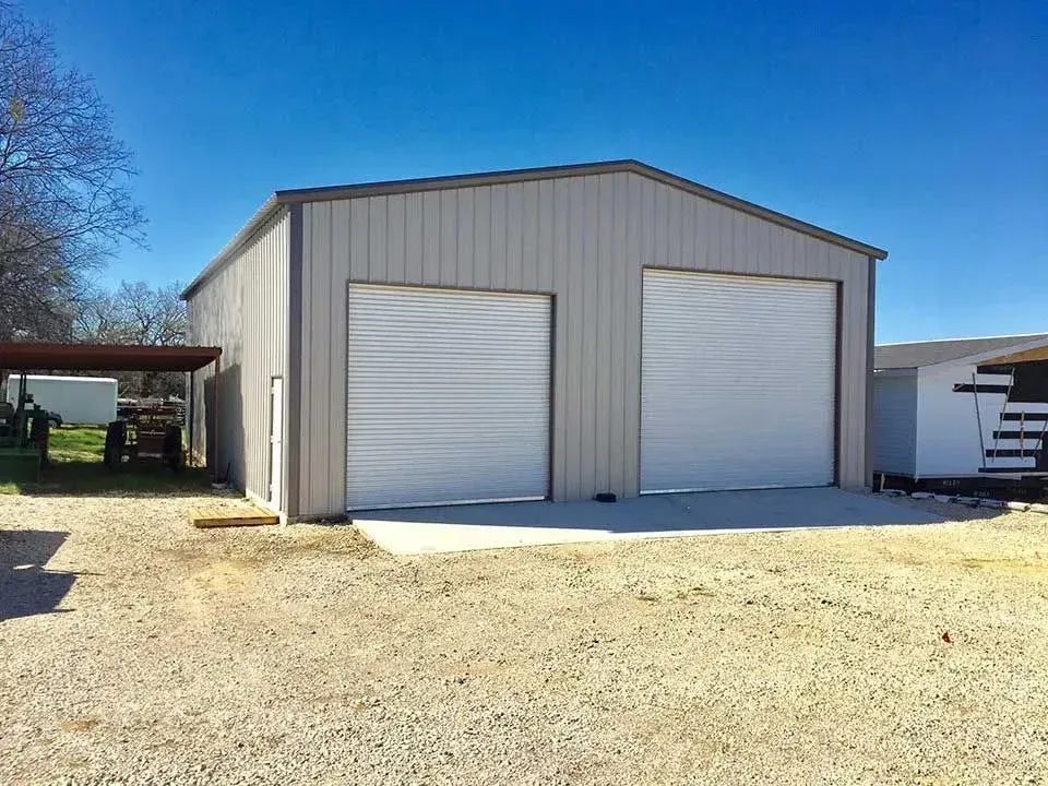 A modern, light-tan metal garage with two large white roll-up doors, set on a gravel lot under a clear blue sky.