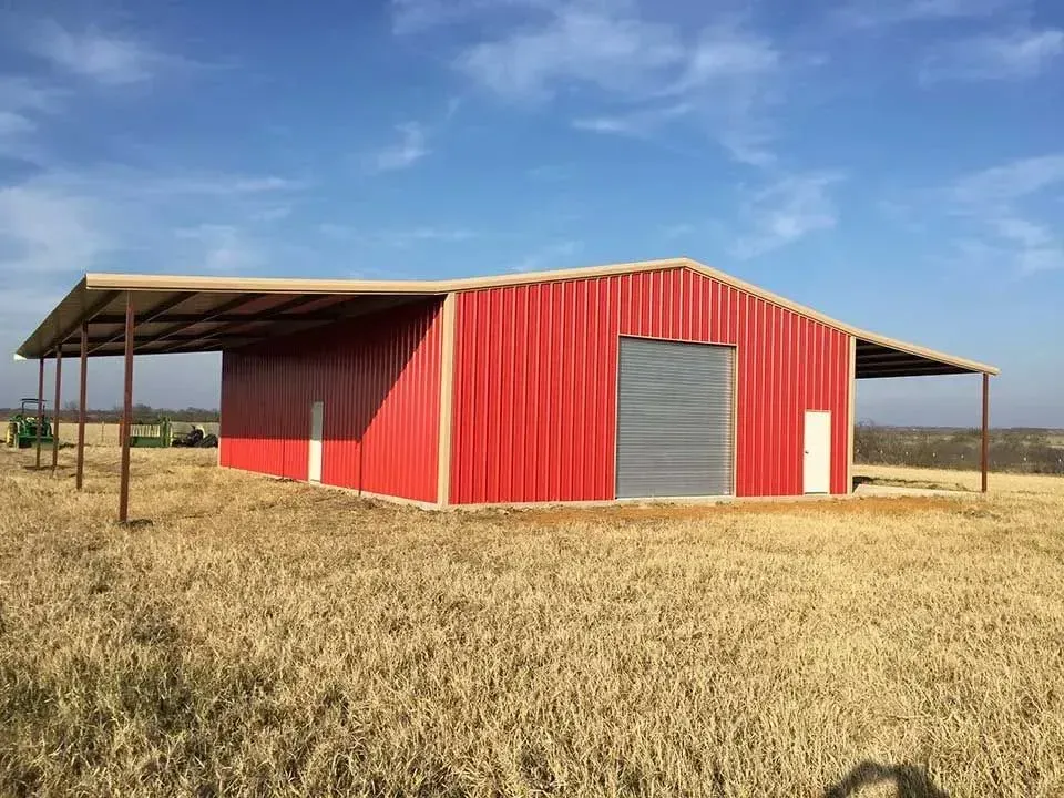A red metal barn with large roll-up doors and side lean-tos sits in an open, grassy field under a blue sky.