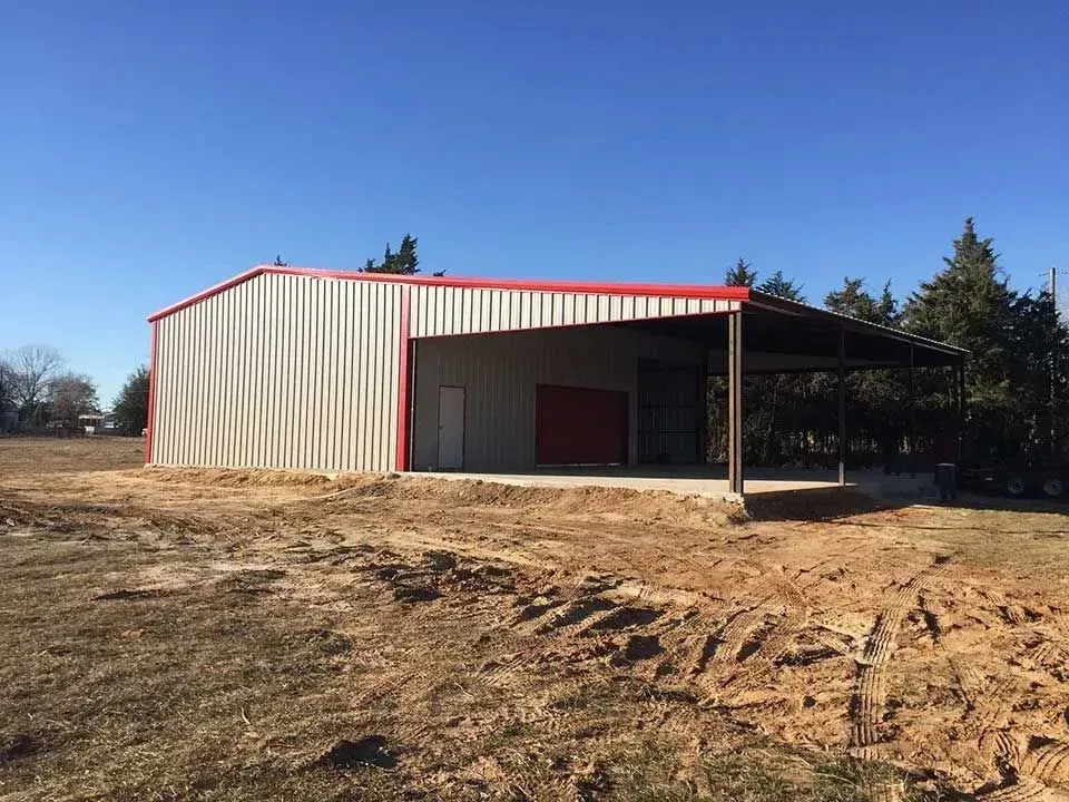A metal utility building with a beige exterior and red trim stands on a dirt lot under a clear, bright blue sky.