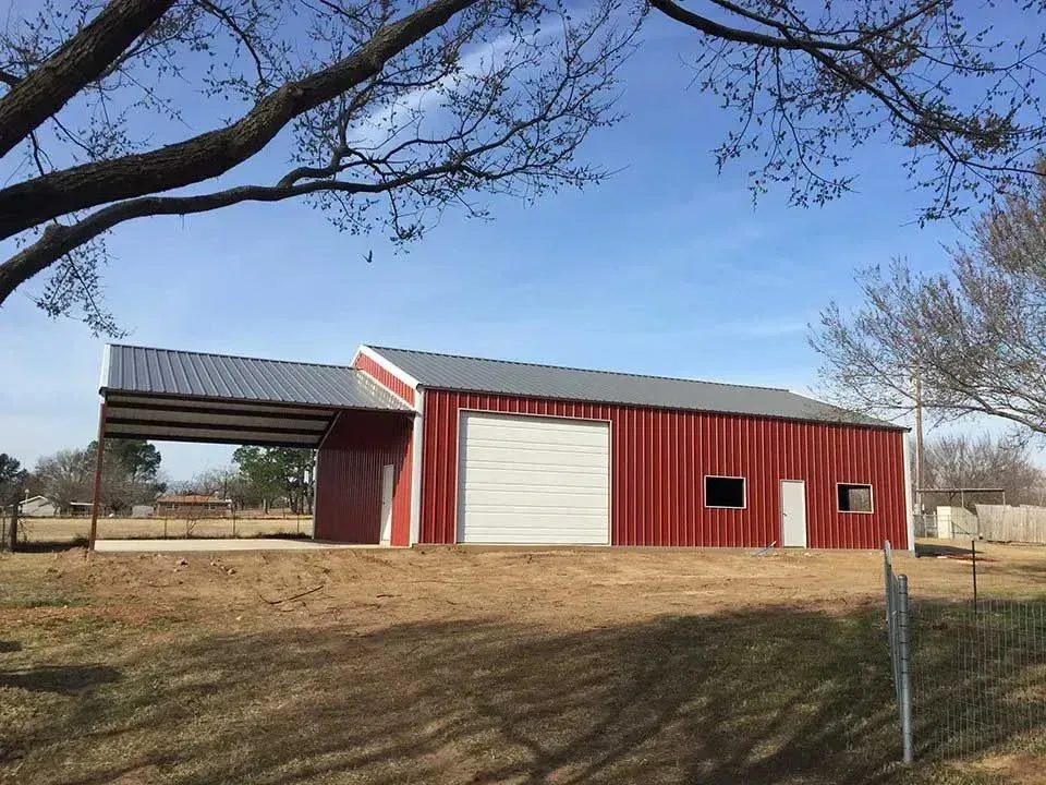 A red metal barn with a white roll-up door and an attached open-sided carport, set on a grassy lot under a blue sky.