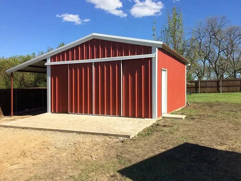 A red metal barn with sliding doors and an adjacent open-sided carport structure on a dirt lot under a blue sky.