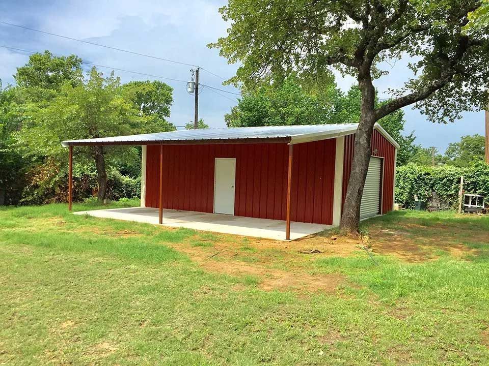 A red metal storage building with a concrete porch and overhang, situated in a green grassy yard near a tree.