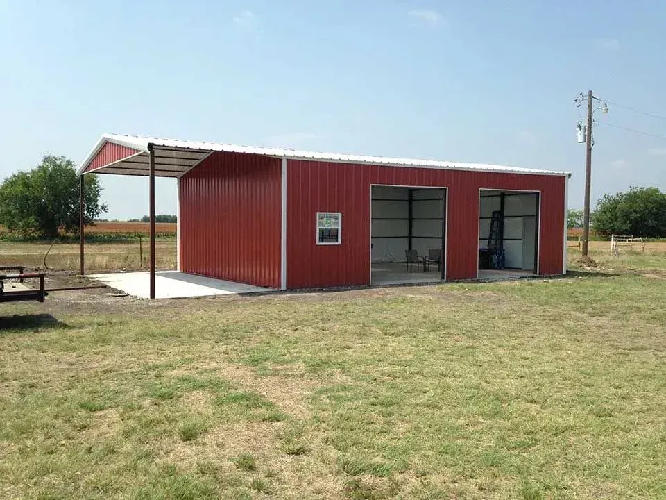 A red metal building with an attached carport sitting in a grassy field under a clear blue sky.