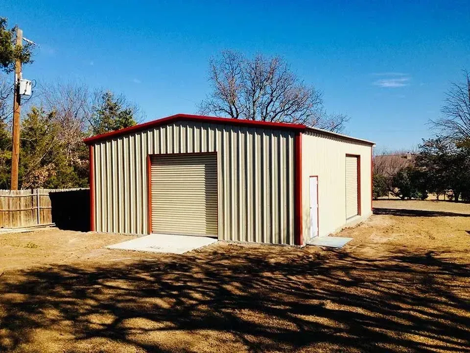 A beige metal workshop with a red-trimmed roof, a roll-up garage door, and a side entry door in a grassy lot.