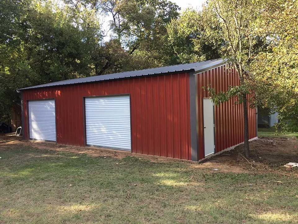 A red metal workshop with two white roll-up garage doors and a side entry door, situated in a grassy, wooded area.