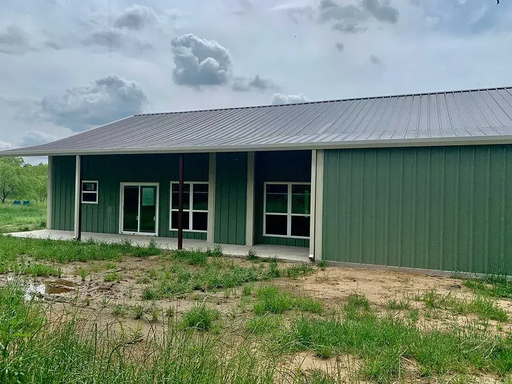 A single-story, green metal building with a grey roof, sliding glass doors, and large windows, set in a grassy field.