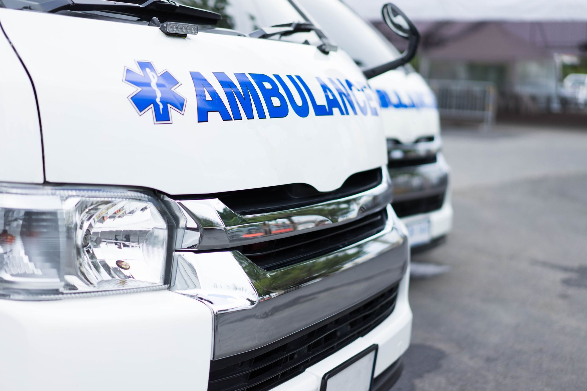 Parte anteriore di un'ambulanza bianca con scritte blu, parcheggiata in strada vicino ad altri veicoli.