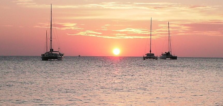 Three sailboats are floating in the ocean at sunset