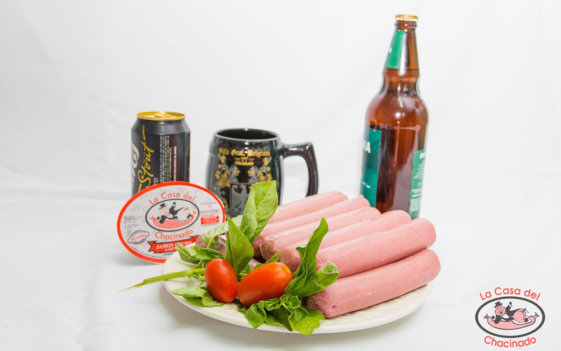 Mesa con salchichas rosas, tomates, lechuga, latas de cerveza, taza y botella sobre un fondo blanco.