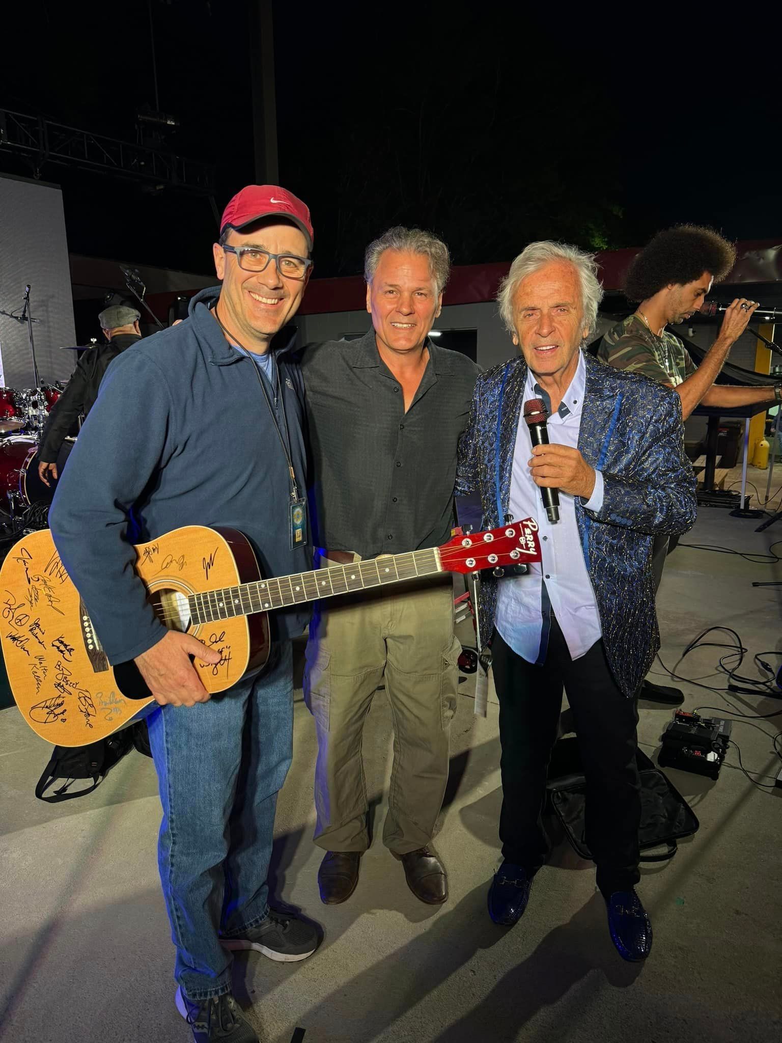 Three men pose for a photo with a guitar and microphone on an outdoor stage at night.