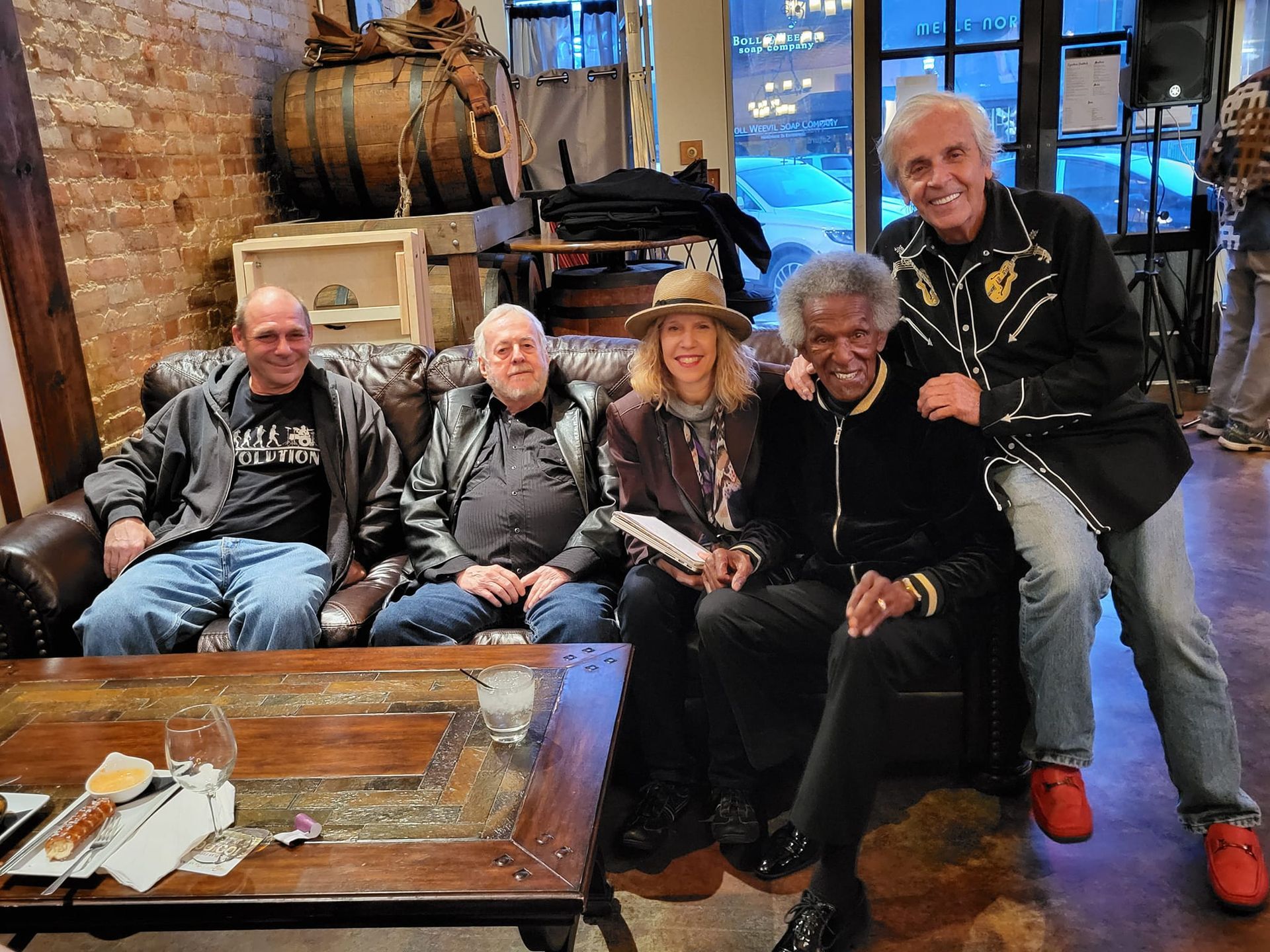 Five people pose on a leather sofa indoors. Dark wood and brick interior. One man leans in from the right.