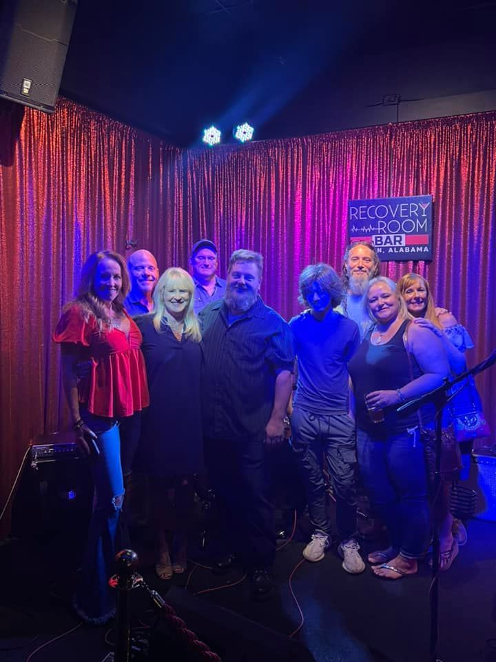 Group of people smiling, posing on a stage with red curtains. Stage is lit with blue and white lights.