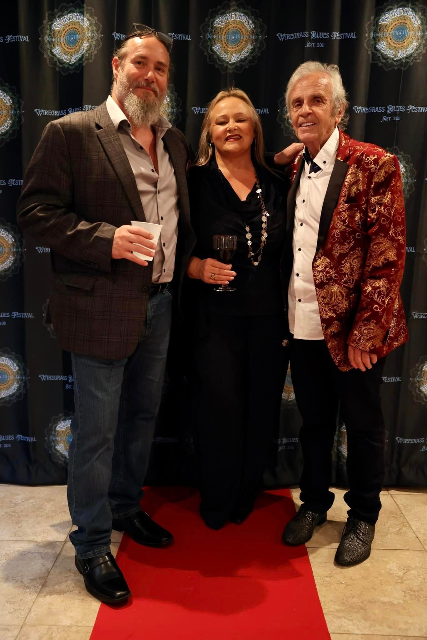Three people pose on a red carpet. A man in a maroon jacket, and two others in formal attire.
