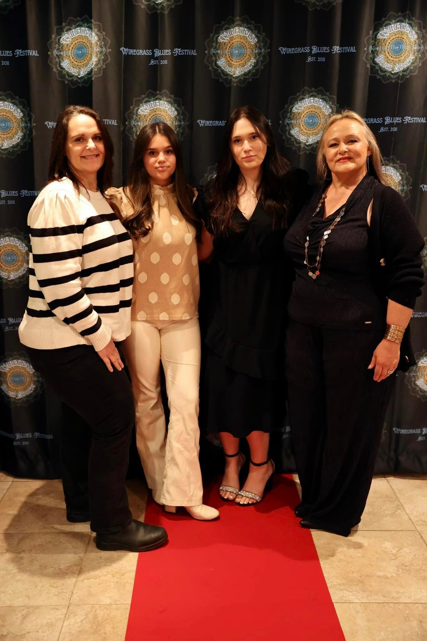 Four people pose on a red carpet in front of a backdrop, two women on each end and two in the middle, smiling.