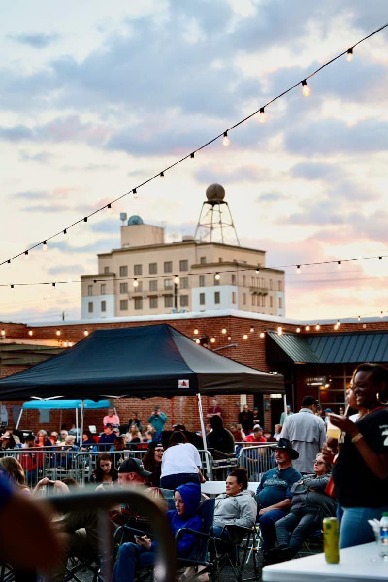 Outdoor gathering under string lights with a brick building and a tall building in the background. People seated, socializing.