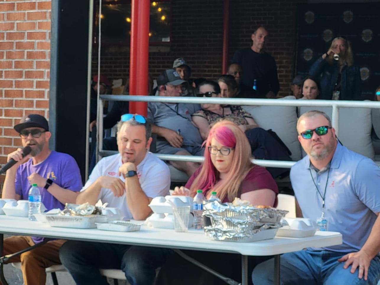 Four people at a table eating food, being judged by two others. Outdoors with onlookers.