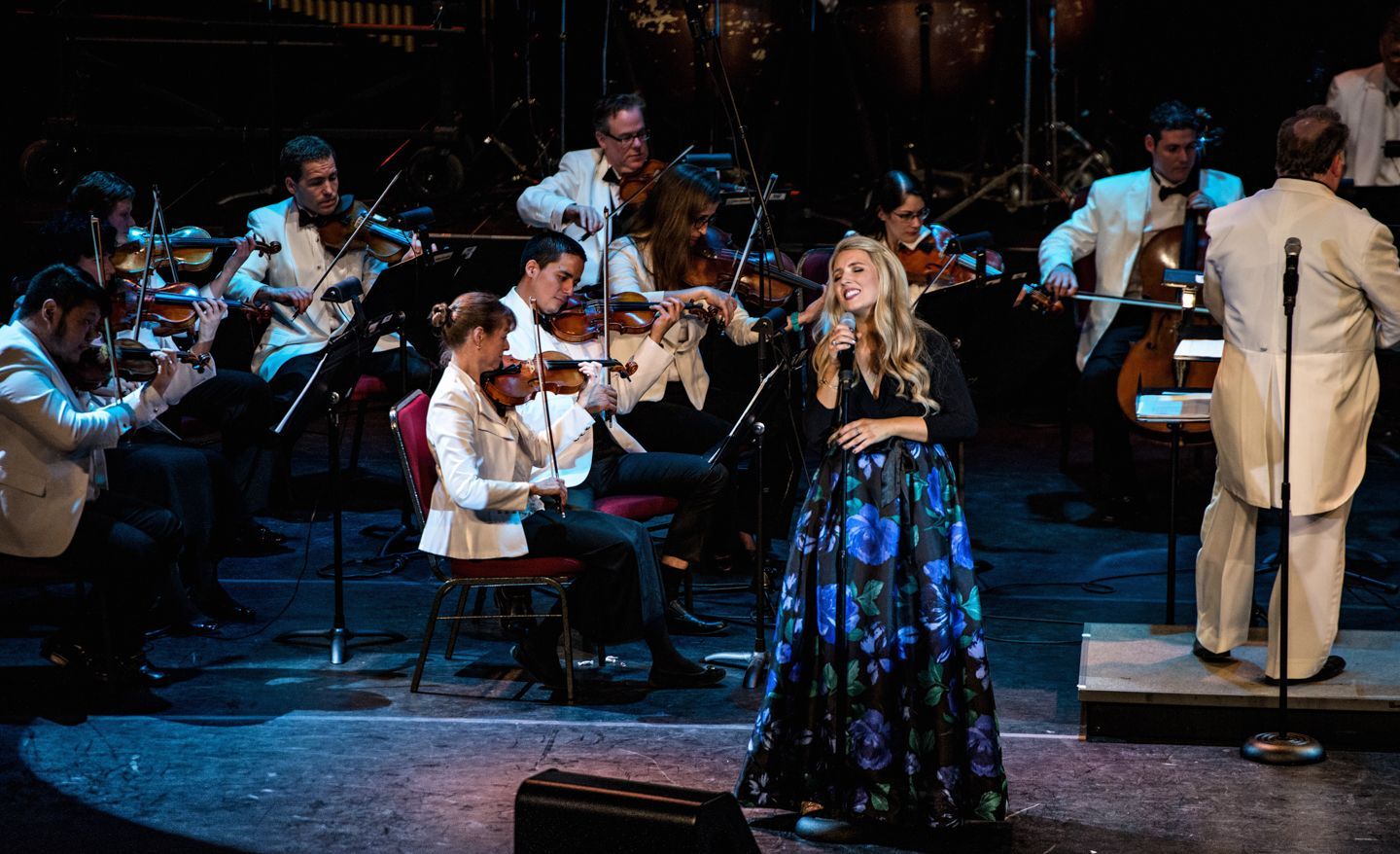 Woman in floral skirt sings with orchestra on stage.