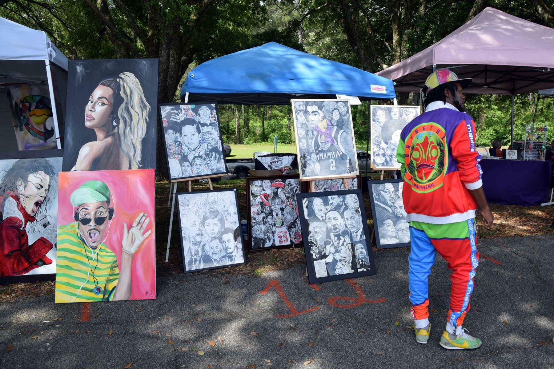 Art display at an outdoor market. Person in colorful outfit looks at art. Paintings of people and faces.
