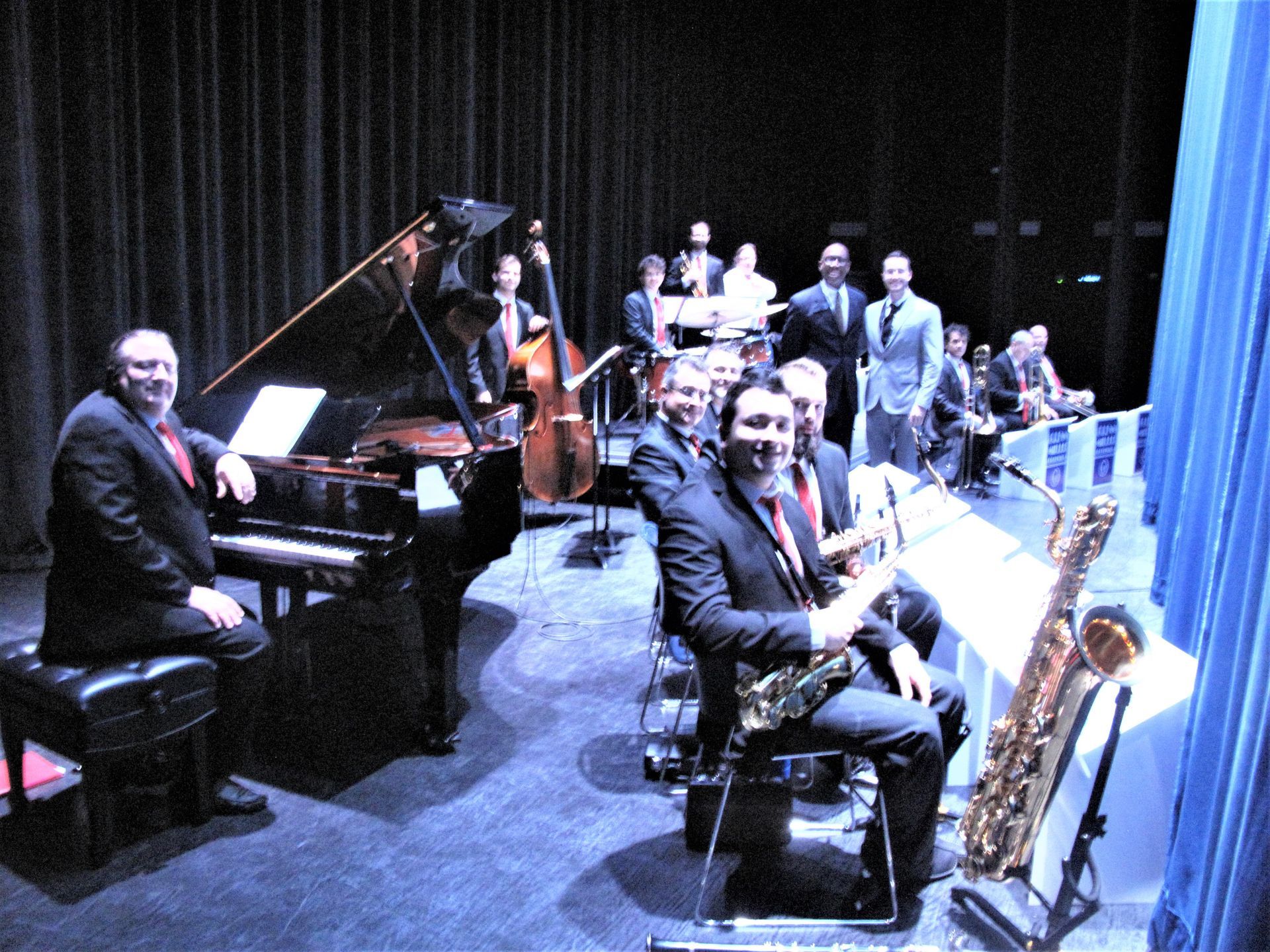 Jazz band on stage, piano player seated, saxophonist in front, other musicians behind, stage lit, blue curtains.