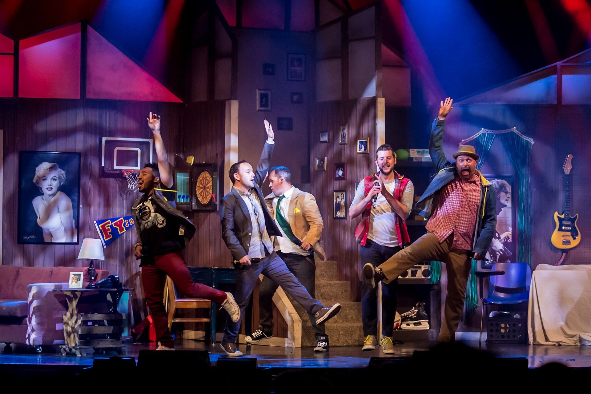 Five men on stage, dressed in various outfits, pose with arms raised. Stage set depicts a room, lights.