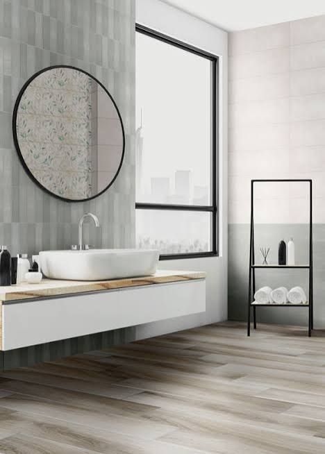 Bagno moderno con lavabo a parete, specchio rotondo, finestra e mensola nera sopra il pavimento effetto legno.