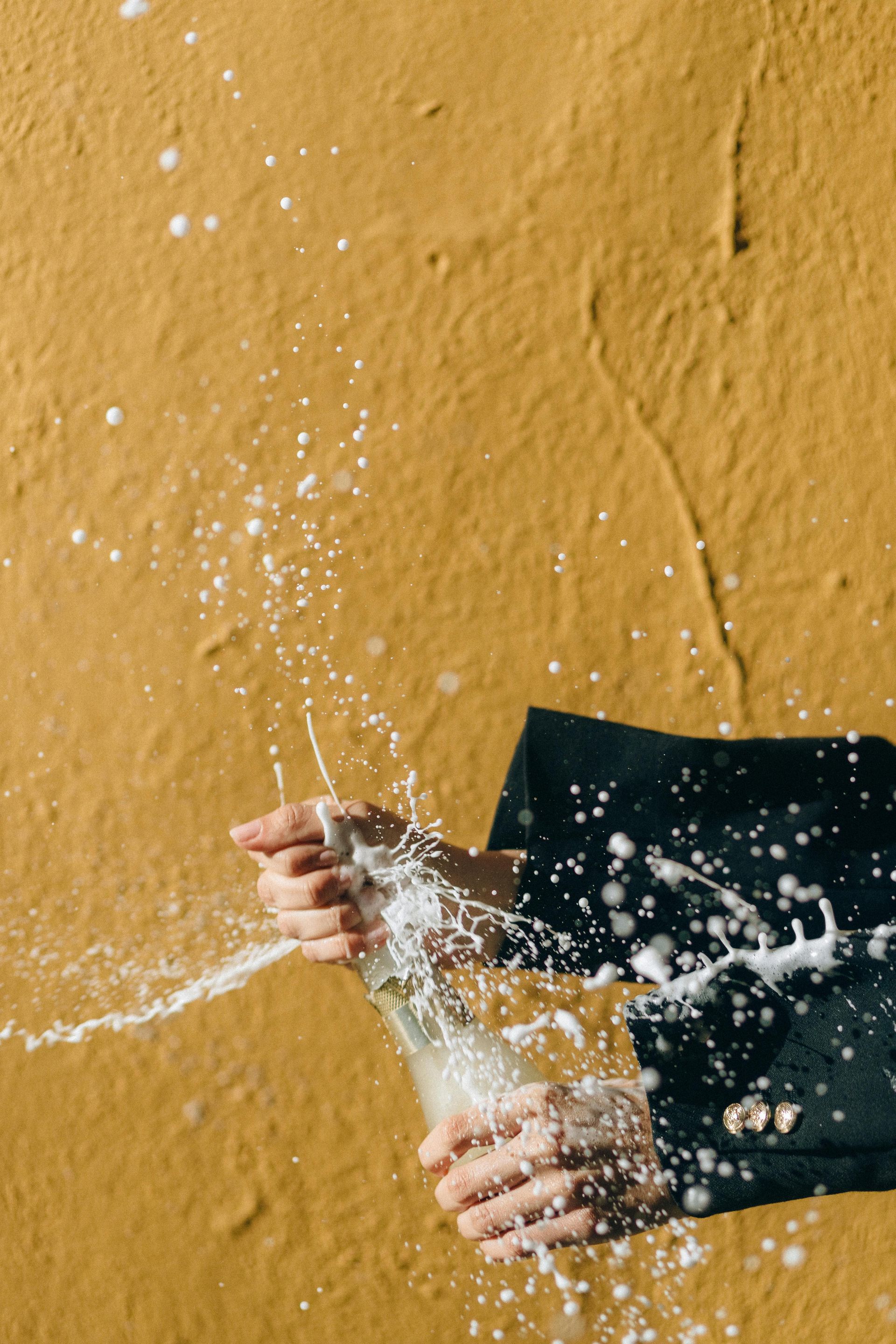 Hands opening champagne bottle, spraying foam against a yellow wall.