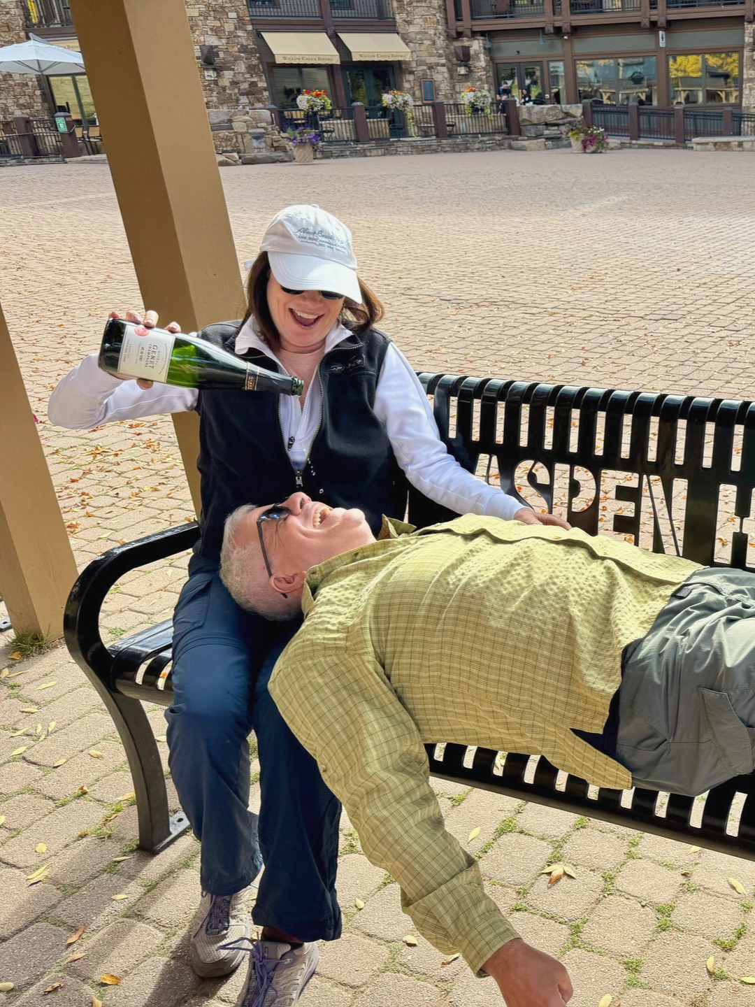 Woman pours liquid from bottle onto man's face while sitting on a bench that reads