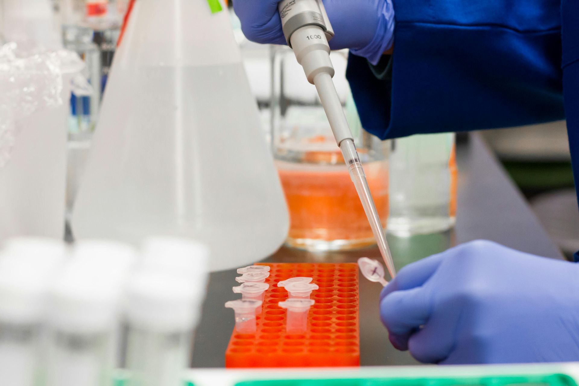 A gloved hand uses a pipette to transfer liquid into a small vial in a laboratory setting.