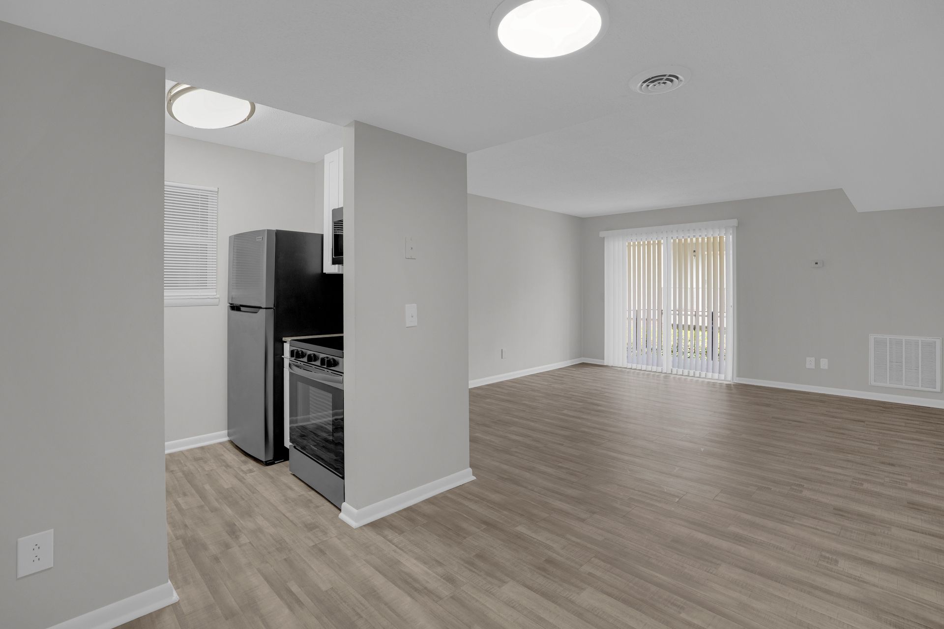 A brightly lit, empty apartment featuring a small kitchen with stainless steel appliances and light wood-tone flooring.