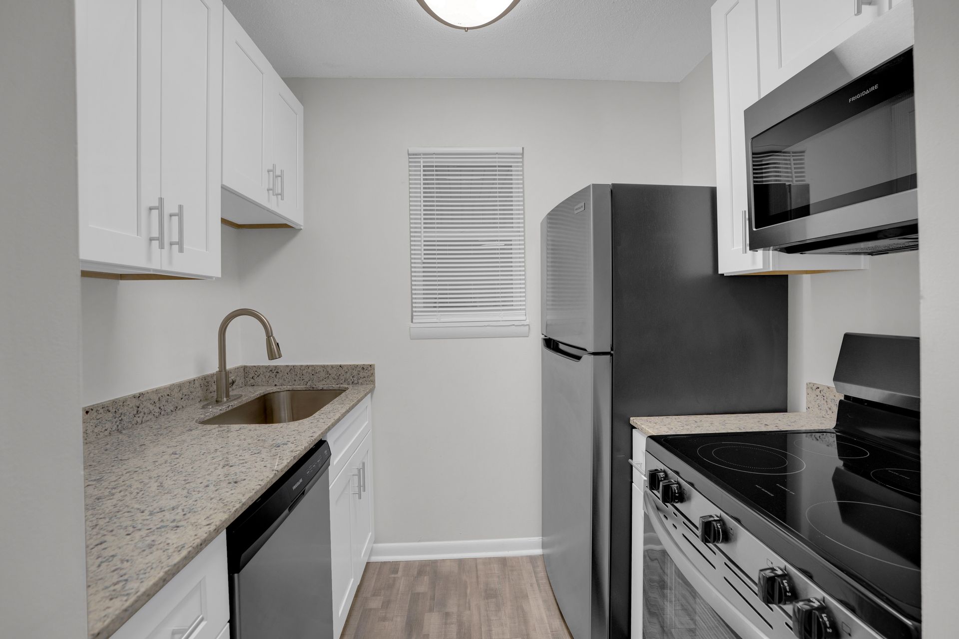 A bright, galley-style kitchen with white cabinets, speckled granite countertops, a stainless steel fridge, and oven.