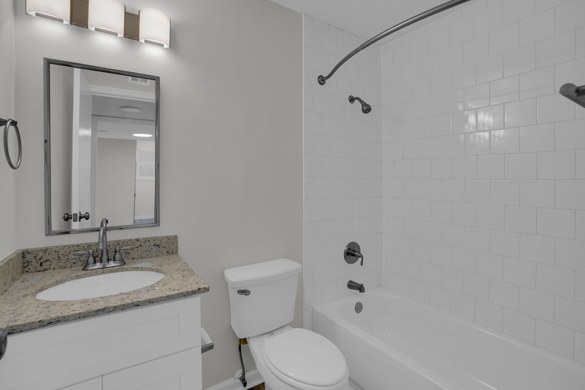 A bright, modern bathroom with a white vanity, a granite countertop, a toilet, and a white tiled shower-tub combination.