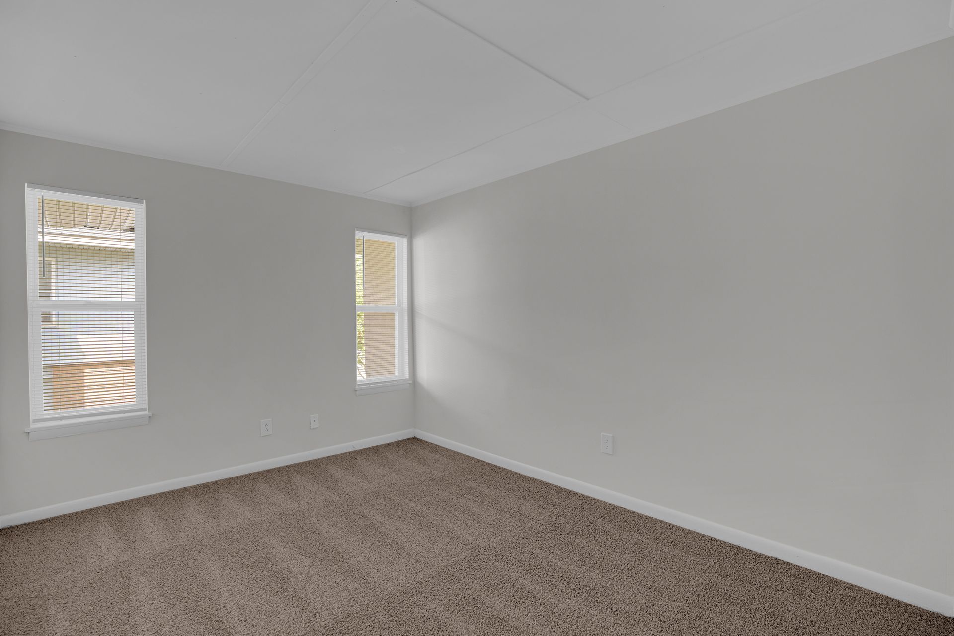 An empty room with grey walls, beige carpet, and two windows on the left wall.