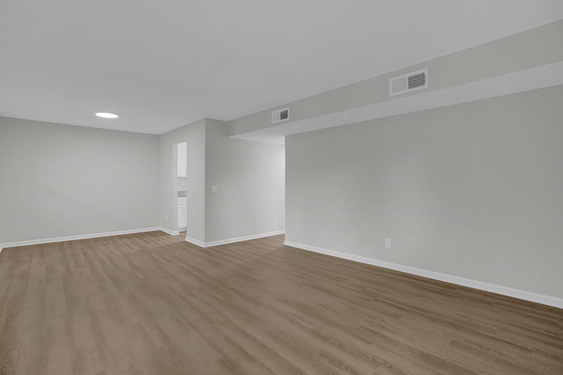 An empty room with light gray walls, brown wood-look flooring, and a recessed ceiling area with two air vents.