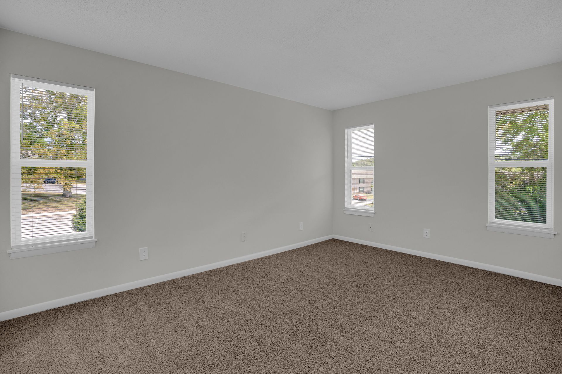 An empty bedroom with gray walls, brown carpet, and three windows letting in natural light.